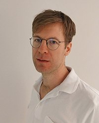 Porträtt Jacob Nissen, vit t-tröja, kort hår och glasögon.